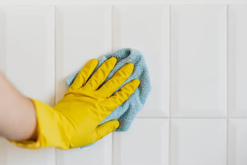 Gloved hand wiping white wall tiles with a microfibre cloth